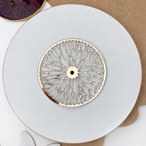 May include: A circular white coaster featuring a centered gold-toned metal inlay with an intricate, radiating petal-like cutout pattern. The design consists of numerous elongated, organic shapes arranged in a sunburst formation around a small center hole.