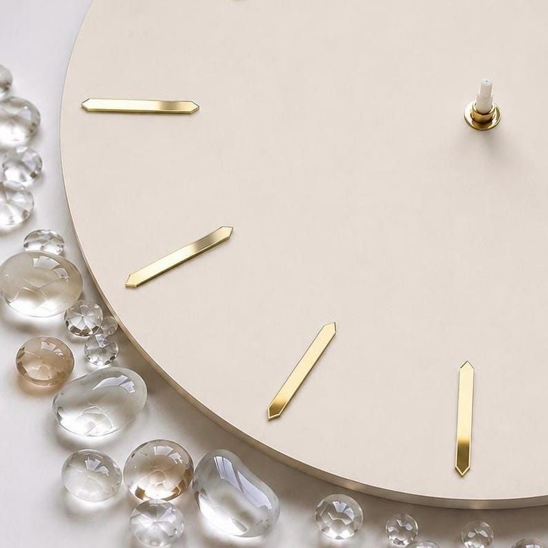 May include: A circular wall clock face in a matte cream finish featuring four gold-toned metal hour markers positioned at intervals. The center mechanism post is visible, and the edge is adorned with a cluster of clear, smooth glass stones.