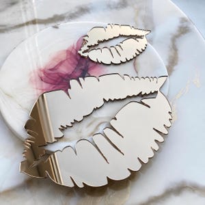 May include: Two reflective, silver-toned lip-shaped cutouts with jagged, textured edges. The smaller lip shape is positioned above the larger lip shape, both featuring a smooth, mirror-like surface that reflects the surrounding environment clearly.