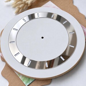 May include: A circular clock face featuring a matte white center with a central hole for a mechanism. A metallic silver ring surrounds the center, accented with twelve evenly spaced rectangular white markers representing the hour positions.