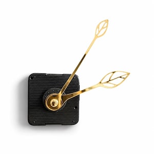 May include: A black square clock mechanism featuring a ribbed surface texture and two gold-colored clock hands. The hands are designed with an openwork leaf-shaped pattern at the ends, extending outward from a central circular gold cap.