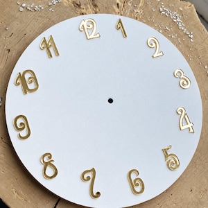 May include: A circular white clock face featuring twelve individual gold-colored numerals arranged in a standard clock layout. Each numeral displays a decorative, swirling font style. A small, centered circular hole is present for a clock mechanism.