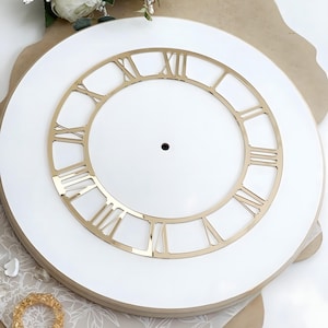 May include: A circular white clock face featuring a metallic gold-colored ring with cut-out Roman numerals. The center of the clock face has a small circular hole for a mechanism. The design is clean and features a smooth, flat surface finish.