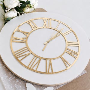 May include: A round wall clock featuring a smooth white circular face and a metallic gold frame with cut-out Roman numerals. The clock has three thin gold hands at the center. A cluster of white artificial flowers sits at the top edge.