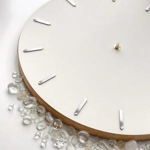 May include: A circular wall clock with a smooth white face and silver-toned rectangular hour markers. The clock features a light-colored wooden edge and a central gold-toned pin. A collection of small, clear, polished stones is arranged along the bottom.