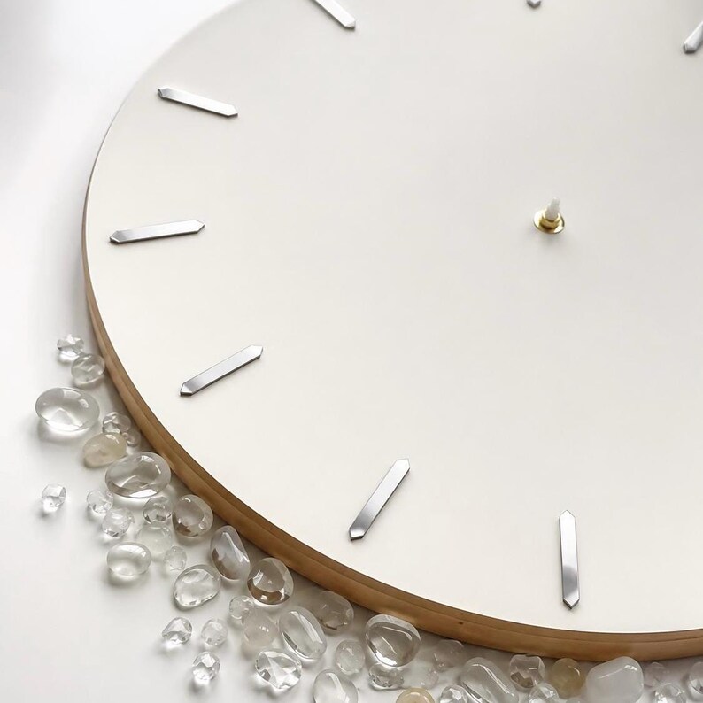 May include: A circular wall clock with a smooth white face and silver-toned rectangular hour markers. The clock features a light-colored wooden edge and a central gold-toned pin. A collection of small, clear, polished stones is arranged along the bottom.