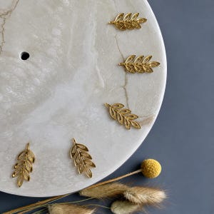 May include: Five gold-toned metal leaf charms arranged in a circular pattern on a smooth, light-colored, marbled circular surface. Each charm features a delicate, open-work branch design with multiple small, pointed leaf shapes along the stem.