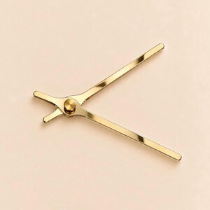 May include: A gold-colored metal clock hand set featuring two long, slender arms and two shorter, stubby arms connected at a central circular pivot point. The surface of the clock hands has a smooth, reflective, and polished metallic finish.