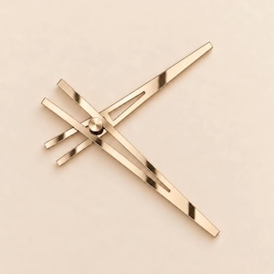 May include: A set of three metallic gold clock hands with a polished finish. The hands are stacked at a central circular pivot point, featuring elongated, tapered shapes with open centers, arranged in a fan-like configuration pointing right.