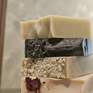 May include: A stack of handmade soaps in various colors and textures. The soaps include a light beige bar, a dark charcoal bar with dried herbs, a bar with oatmeal, and a light pink bar with dried rose petals. The soaps are stacked on top of each other.