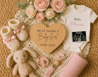 May include: A baby announcement arrangement with a wooden heart reading "We're Having a Baby Girl" and "April 202X". Includes a white babygrow with "Baby Olivia" and an ultrasound image, pink booties, a plush bunny, and a dummy.