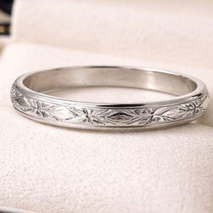 May include: A silver ring with a floral pattern, displayed in a black jewelry box with a gold trim. The ring is centered in the box, showcasing its intricate design. The ring's surface is engraved with a repeating pattern of leaves and flowers.