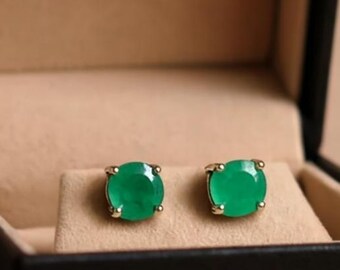 Puede incluir: Un par de pendientes de botón verde esmeralda expuestos en una caja de joyería negra abierta con interior beige. Los pendientes son redondos y están engastados en metal dorado.