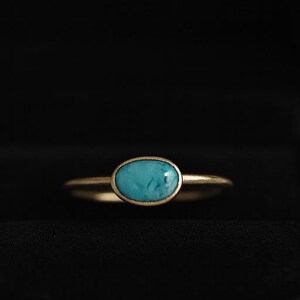 Könnte beinhalten: Ein goldener Ring mit einem ovalen türkisfarbenen Stein, präsentiert in einer offenen schwarzen Samt-Schmuckschatulle. Der Ring ist vor dunklem Hintergrund zentriert, wodurch Design und Farbe hervorgehoben werden.
