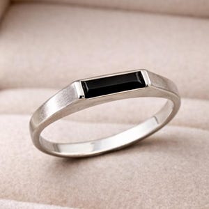 May include: A silver ring featuring a rectangular black gemstone. The ring has a brushed finish and is presented in a beige ring box. The design is minimalist and contemporary, suitable for everyday wear.