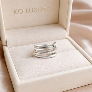 May include: A silver ring with a unique design, featuring three stacked bands and a cylindrical accent. The ring is displayed in a cream-colored jewelry box with the text "KO LUXURY" embossed on the lid.