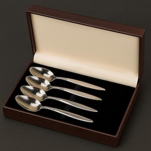 Könnte beinhalten: Ein Set aus vier silbernen Löffeln, präsentiert in einer braunen Präsentationsbox. Die Löffel haben ein dekoratives Design am Griff und liegen auf einer schwarzen, samtartigen Oberfläche.