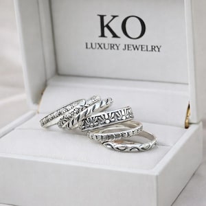 Puede incluir: Una colección de anillos de plata con varios diseños florales y estampados, expuestos dentro de una caja de joyería blanca. La tapa de la caja está abierta, revelando los anillos. El texto "KO LUXURY JEWELRY" está impreso en el interior de la caja.