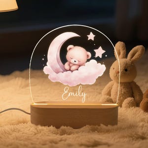 May include: A night light featuring a sleeping teddy bear on a crescent moon and cloud design. The light is made of clear acrylic and sits on a wooden base. The name "Emily" is written in script. A plush bunny is in the background.