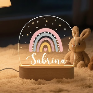 May include: A personalized night light featuring a rainbow design in pastel colors, with the name "Sabrina" in a script font. The light sits on a wooden base and is accompanied by a stuffed bunny.