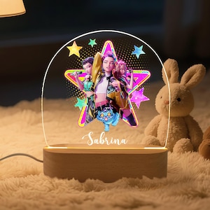 May include: A decorative night light with a wooden base and a clear acrylic top, displaying a colourful graphic of three figures within a star shape. The name "Sabrina" is written below. A soft glow emanates from the light. A stuffed bunny is in the background.