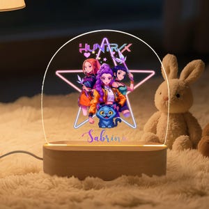 Könnte beinhalten: Dekorative LED-Nachtlampe mit einer Cartoon-Illustration von drei Mädchen und einer blauen Katze, mit dem Text "HUNTRX" und "Sabrina". Das Licht steht auf einem Holzsockel und neben einem Stoffhasen.