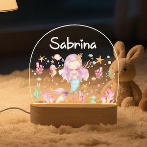 May include: A personalized night light featuring a mermaid design with the name "Sabrina" in white script. The acrylic panel sits on a wooden base, and the design includes sea creatures and coral in pastel colors.