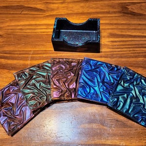 May include: A set of iridescent coasters in shades of purple, green, bronze, and blue, arranged in a semi-circle. A matching black business card holder sits above the coasters on a wooden surface.