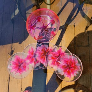 May include: A set of clear glass coasters and a matching glass, all featuring a vibrant pink floral design. The coasters are round, and the glass has a similar pattern. The items are arranged on a glass table.