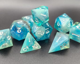 May include: A set of polyhedral dice in shades of translucent blue and teal with swirling white inclusions. Each die features sharp edges and clear, silver-colored engraved numbers. The collection includes various geometric shapes used for gaming.