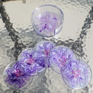 May include: A set of six clear resin coasters with a floral design in shades of purple and pink. The coasters are arranged around a matching holder, also clear with a purple floral pattern. The set is designed to protect surfaces from drink rings.