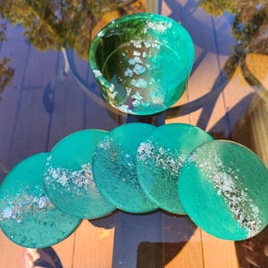 May include: A set of six teal resin coasters with a matching coaster holder. The coasters and holder have a marbled effect with silver accents. The coasters are round and approximately 4 inches in diameter.