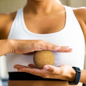 May include: A light brown massage ball is held between two hands. The person is wearing a white sports bra and a black watch. The ball appears to be made of cork or a similar material, and is used for muscle therapy.