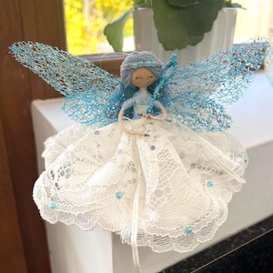 May include: A decorative fairy figurine with light blue yarn hair, a white lace dress, and shimmering blue mesh wings. The figure features a painted face, a blue beaded headband, and holds a thin blue beaded wand, accented with small blue beads.