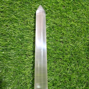May include: A clear, cylindrical crystal wand with a pointed tip. The object features a smooth, polished surface with subtle internal striations and a translucent appearance, standing vertically as a single, solid, elongated mineral formation.