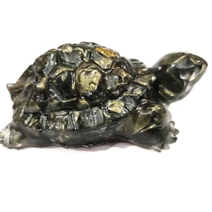 May include: A small, carved turtle figurine made of a dark, polished stone with metallic gold-colored inclusions throughout its shell and head. The surface is smooth and reflective, showing a detailed, textured pattern on the shell.