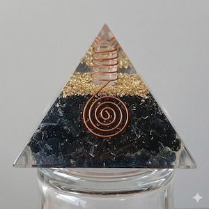 May include: A clear resin pyramid containing a central quartz crystal wrapped in copper wire, a layer of gold-colored metallic flakes, and a base layer of dark black stone fragments. A copper wire spiral is embedded on the front face of the pyramid.