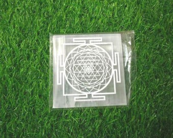 Puede incluir: Una placa cuadrada de selenita blanca translúcida con un patrón geométrico Sri Yantra grabado en blanco. La placa está encerrada en una funda protectora de plástico transparente, mostrando una compleja disposición de triángulos y pétalos entrelazados.