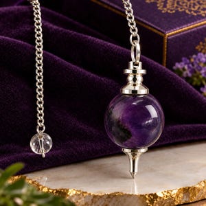 May include: A pendulum featuring a spherical purple amethyst stone with dark inclusions, set between a polished silver-toned metal cap and a pointed silver-toned metal tip. The pendulum hangs from a silver-toned metal chain with a small clear bead.
