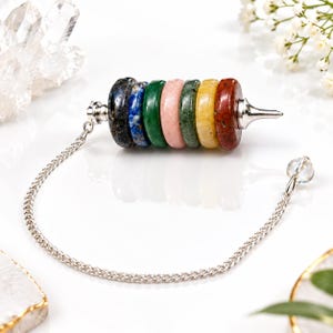 May include: A pendulum featuring seven stacked, circular gemstone beads in black, blue, green, pink, dark green, yellow, and red. The beads are held by a silver-toned metal frame with a pointed tip and a silver chain ending in a clear crystal bead.