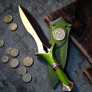 Puede incluir: Una daga de doble filo con hoja de metal pulido y mango verde brillante con una banda metálica. La daga descansa sobre una funda de cuero verde adornada con un emblema triquetra plateado, rodeada por varias monedas dispersas.