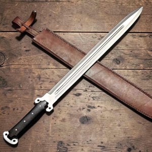 Puede incluir: Una espada plateada con empuñadura negra y funda de cuero marrón. La espada tiene una hoja de doble filo y está sobre una superficie de madera. La funda está cosida y tiene una correa.