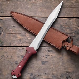 Puede incluir: Una espada de hoja plateada con empuñadura roja y funda de cuero marrón. La espada tiene una punta afilada y una hoja ancha. El mango es de madera y tiene una serie de puntos blancos. La funda tiene una correa y una hebilla.