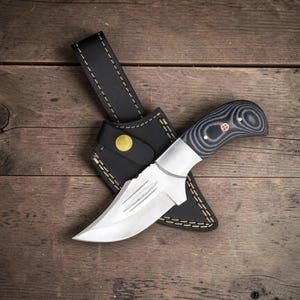 Puede incluir: Un cuchillo de hoja fija con una hoja de acero de color plateado que presenta tres ranuras horizontales y un mango de capas negras y grises con un pasador decorativo. El cuchillo viene con una funda de cuero negro con costuras amarillas y un broche de latón.