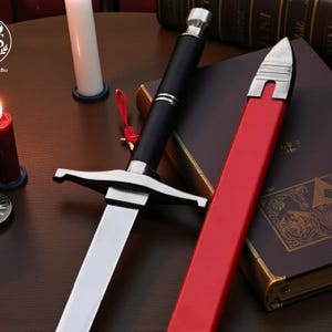 May include: A medieval-style sword with a black handle and silver accents, resting on a wooden surface. A red sheath lies beside it. A lit red candle and a small clock are also visible.