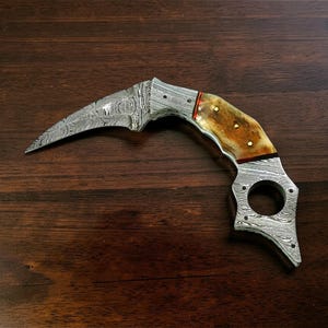 May include: A curved Damascus steel knife with a bone handle. The blade has a distinctive pattern, and the handle features a brown and tan colour scheme with brass accents. The knife has a finger hole at the base of the handle.