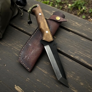May include: A handcrafted knife with a wooden handle and a black blade rests in a brown leather sheath. The sheath features an embossed design and a snap closure. The knife is designed for outdoor use.