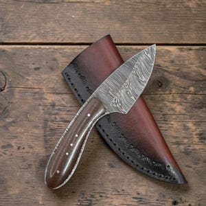 Puede incluir: Un cuchillo de hoja fija con una hoja de acero de Damasco estampada y un mango de madera marrón oscuro asegurado con cuatro pasadores de metal. El cuchillo se coloca sobre una funda de cuero marrón oscuro con costuras visibles.