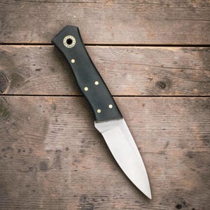 Puede incluir: Un cuchillo de hoja fija con una hoja de acero de color plateado pulido y un mango negro mate. El mango está asegurado con cuatro pequeños remaches de color latón e incluye un orificio decorativo circular para cordón.