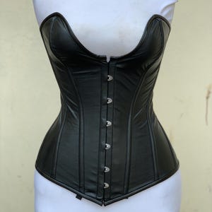 May include: A black corset featuring a sweetheart neckline and a front closure with silver-toned metal busk hooks. The garment is constructed with vertical panels and dark piping along the edges, designed to fit a torso shape with a curved waistline.